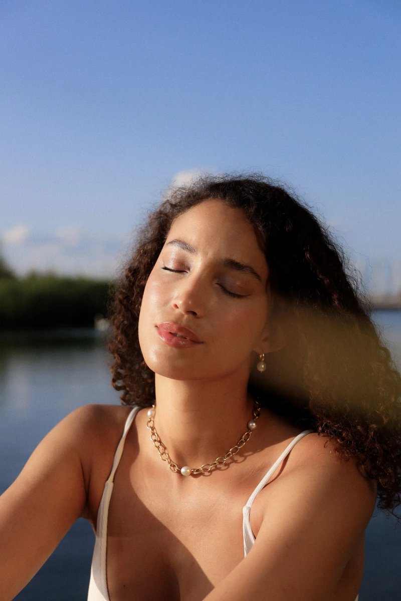 Femme aux cheveux bouclés, yeux fermés, éclairée par une lumière dorée près de l’eau.
