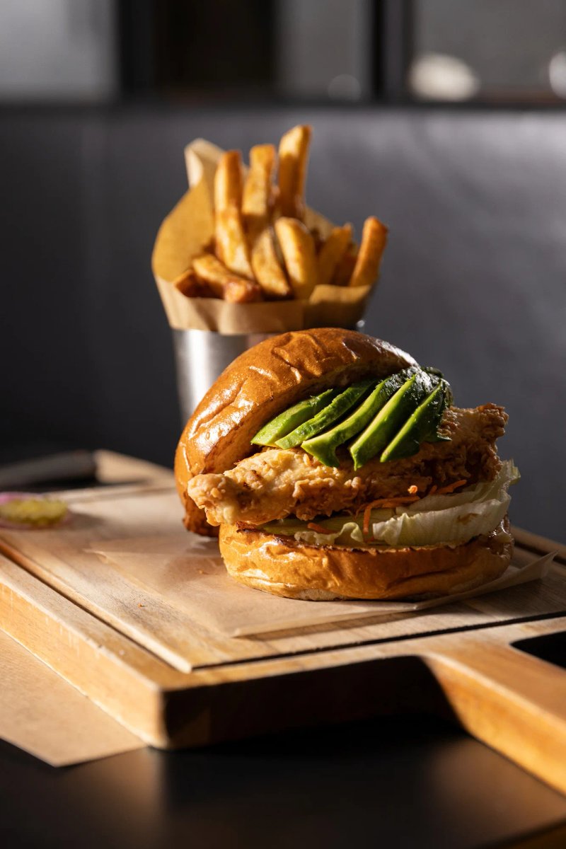 Burger au poulet croustillant et avocat servi avec des frites sur une planche en bois.
