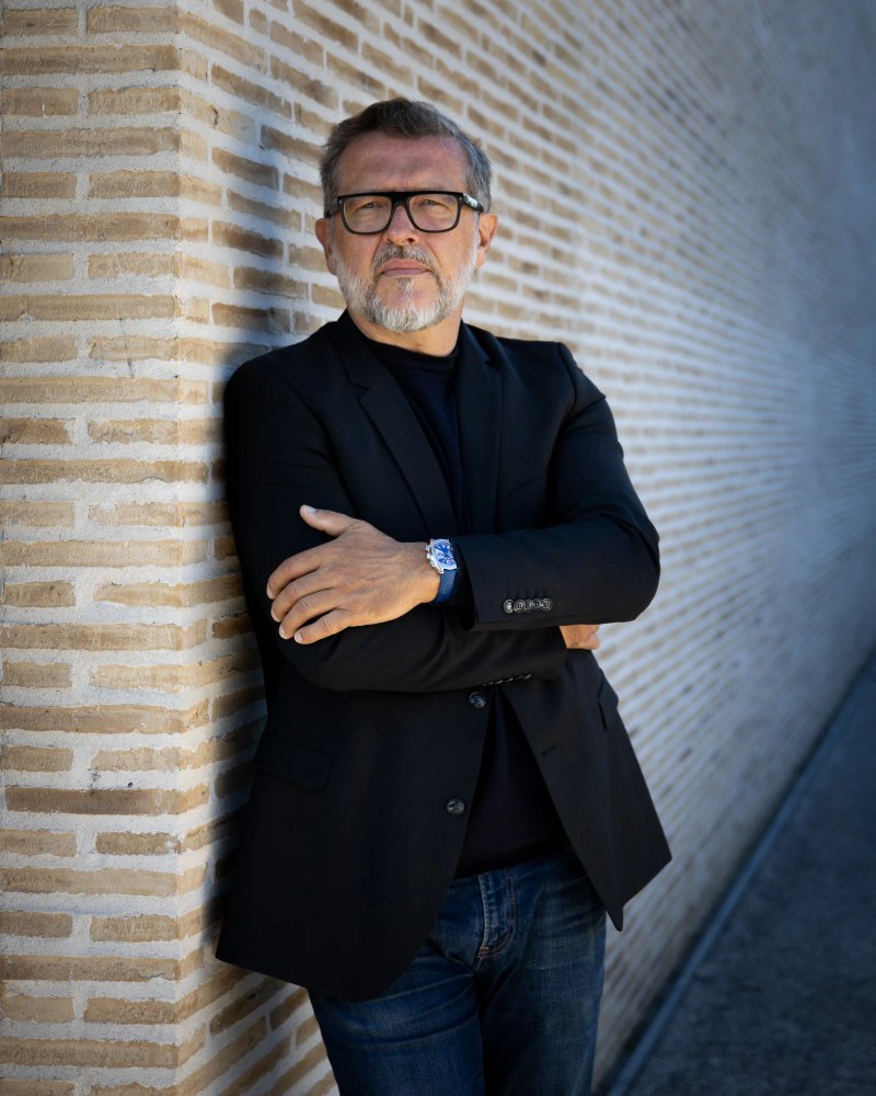 Portrait d’un homme barbu à lunettes, bras croisés devant un mur de briques.