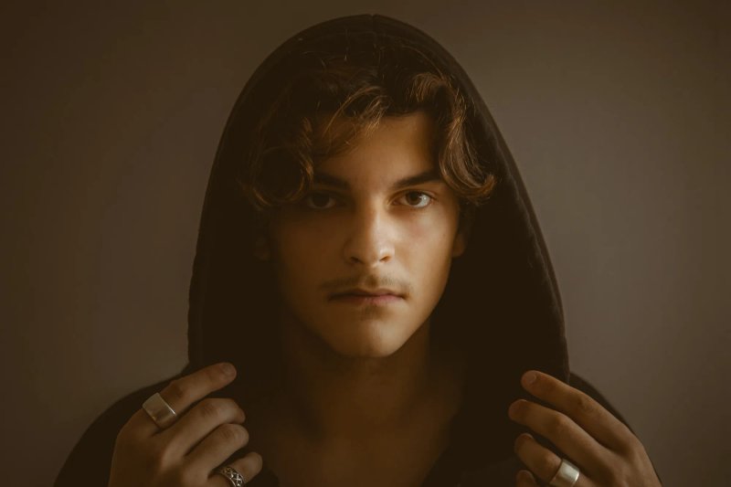Portrait rapproché d’un jeune homme aux cheveux bouclés sous une capuche sombre.