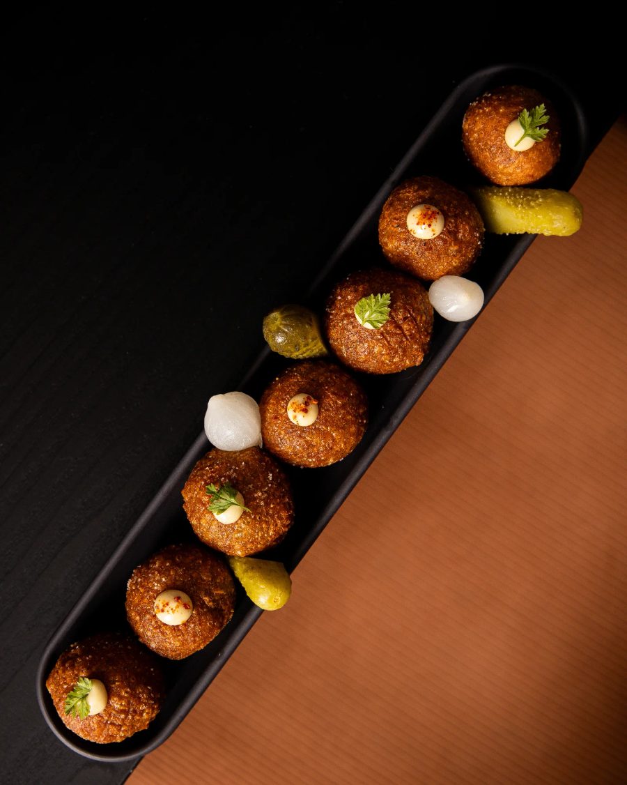 6 boulettes frites disposées en ligne sur plateau noir avec cornichons et oignons, garnies de sauce et persil