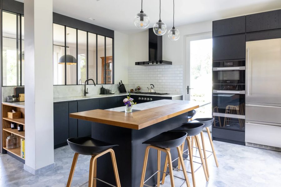 Cuisine moderne noire, îlot central avec plan de travail bois clair et marbre, mur verrière, tabourets design.