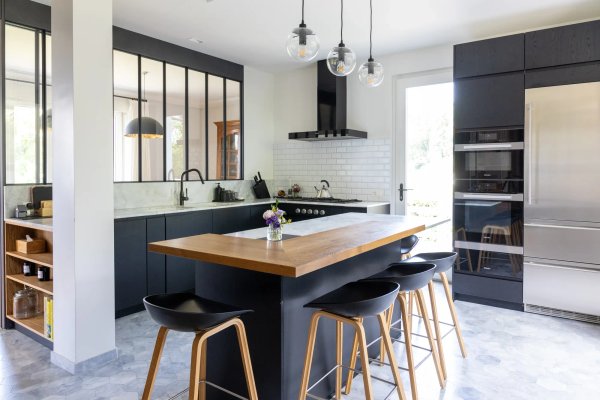 Cuisine moderne noire, îlot central avec plan de travail bois clair et marbre, mur verrière, tabourets design.