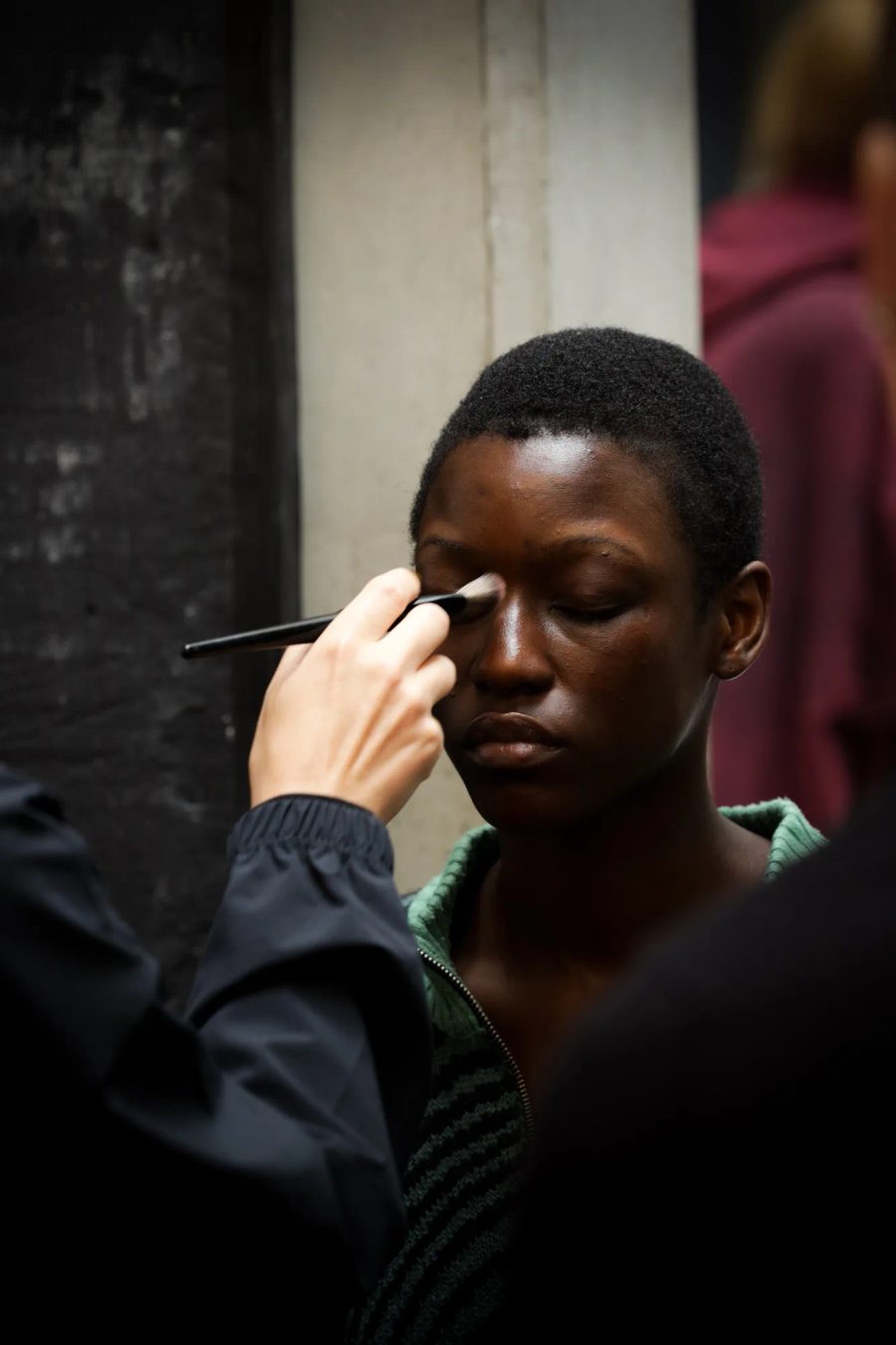 Gros plan : une jeune femme noire aux yeux clos se fait maquiller le visage par une main qui tient un pinceau.
