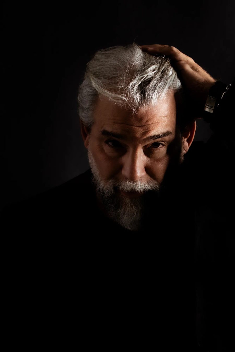 Portrait rapproché d'un homme barbu aux cheveux gris, main sur la tête, regard intense, éclairage dramatique