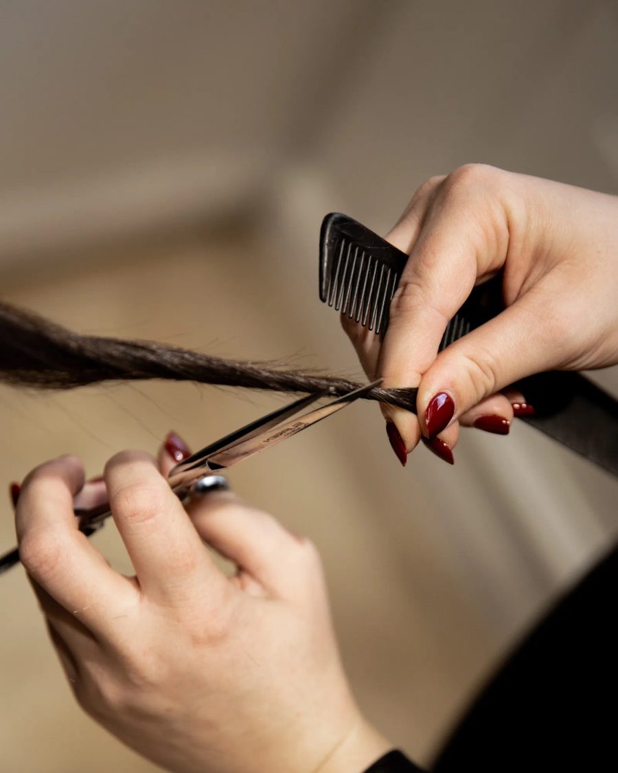 Coiffeuse avec manucure rouge coupe une mèche de cheveux bruns à l'aide de ciseaux et d'un peigne noir
