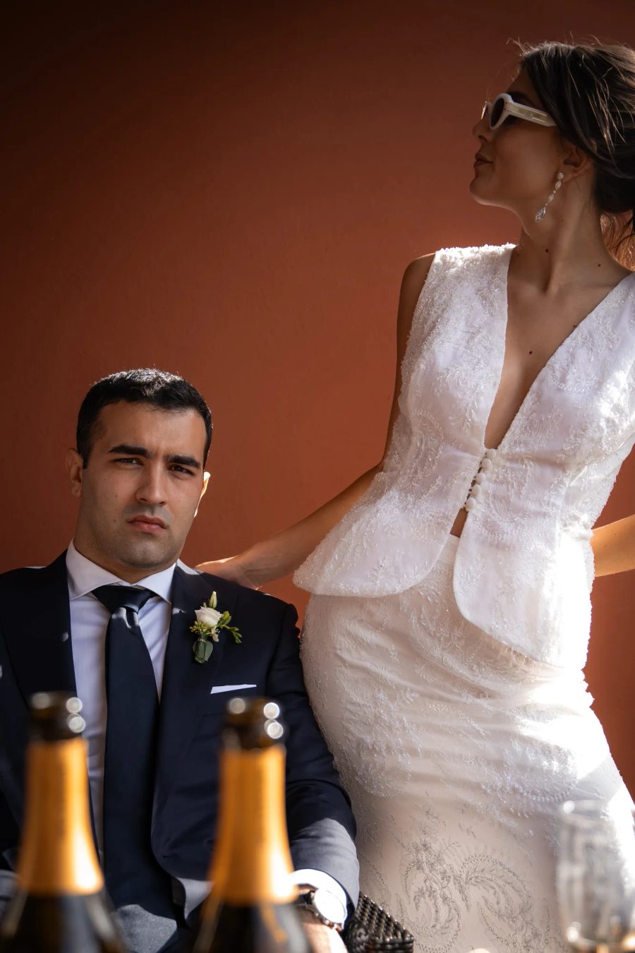 Mariés en tenue formelle, la mariée en robe claire et lunettes de soleil, le marié en costume sombre, avec des bouteilles floues au premier plan.