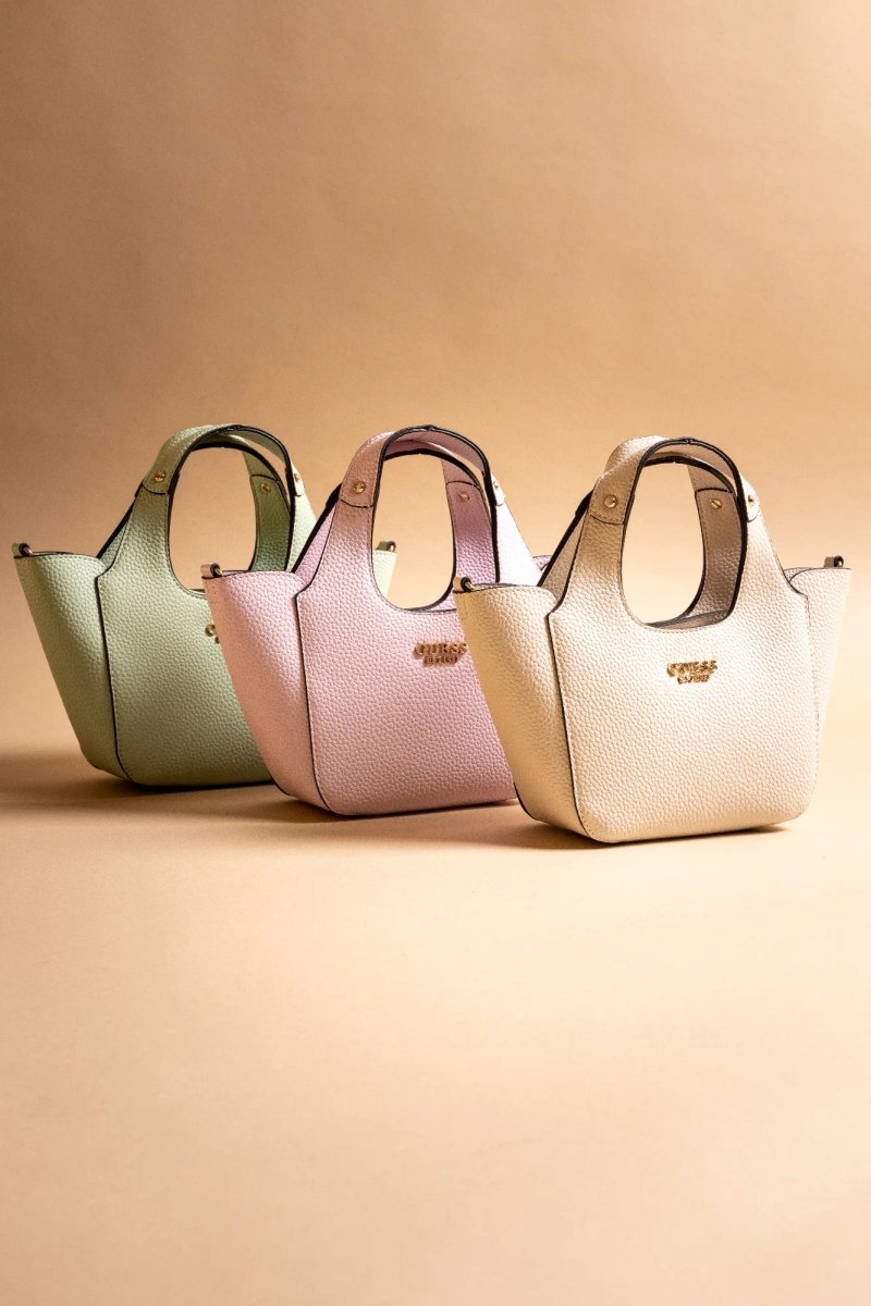 Trois mini sacs Guess pastel, vert menthe, lilas et crème, alignés sur un fond assorti.